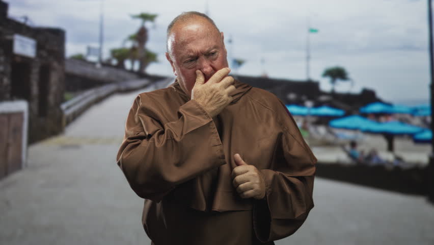 Man monk wearing robe and cross necklace, hand to face touching gesture on street promenade near beach; reflection solitude sorrow.