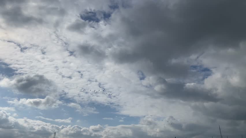 A tranquil winter landscape featuring a soft blue sky with scattered clouds and a distant flock of migrating cormorants.Cormorant Migration on Calm Winter Day.Serene winter sky with sun behind clouds and distant migrating birds.