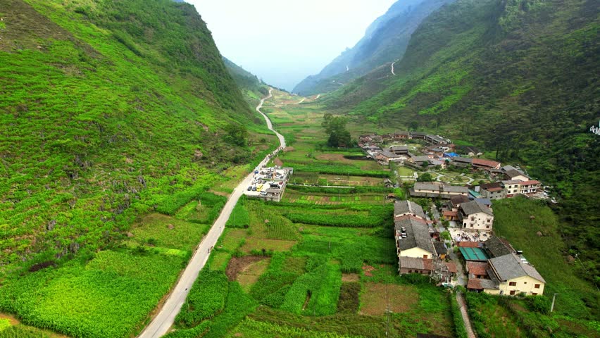 4K aerial pull-back shot from a rock-mounted village sign to reveal the Hmong Pa Vi tourist village nestled at the foot of limestone mountains in Northern Vietnam.
