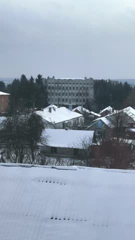 A serene winter landscape captures a quiet village with snow-laden rooftops, a prominent multi-story building, and a backdrop of evergreen trees under a muted, overcast sky. Bare branches frame the vi