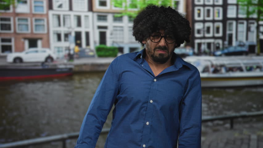 Man grimacing and leaning on a railing beside canal boats on a street in amsterdam; unease hesitation travel.