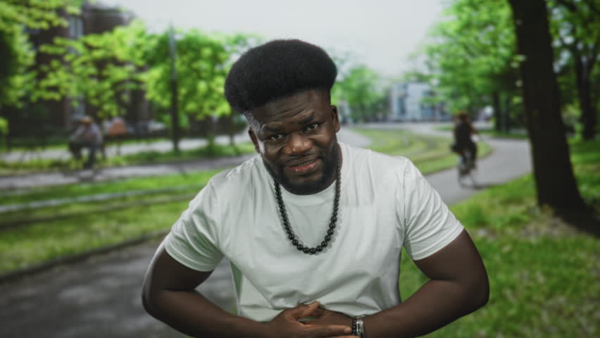 Man holding stomach with beaded necklace on a green street path near trees and tram tracks by buildings; discomfort.