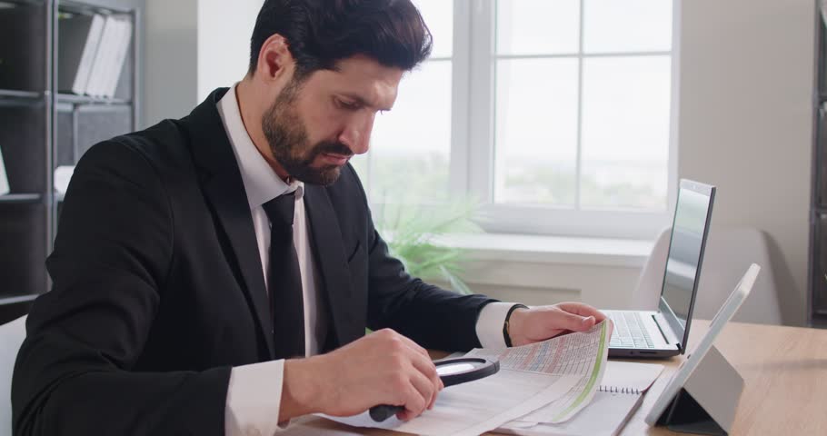 Serious busy man examining paper documents, businessman at desk using magnifying glass, focused, working diligently, detailed scrutiny and investigation, study important contract, sitting at desk