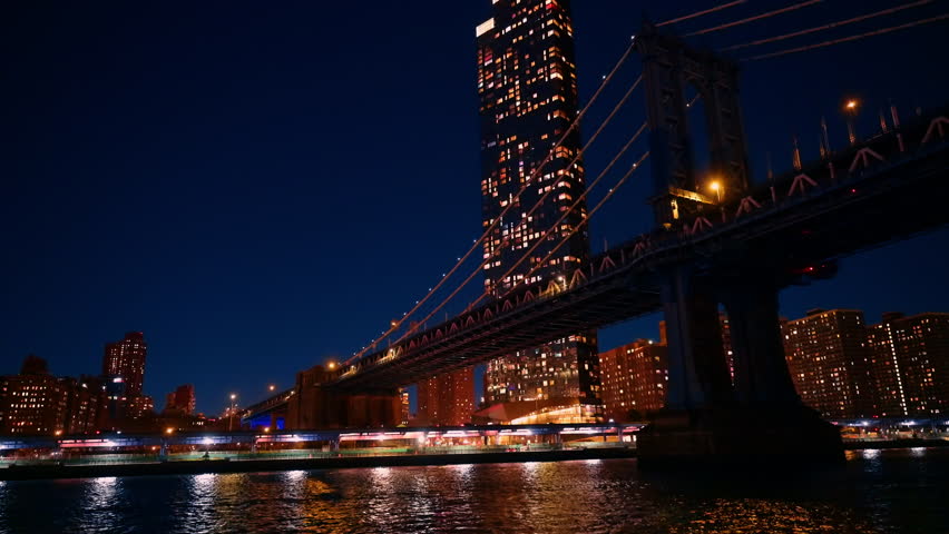 Evening view of a city bridge with buildings and water at night. The city bridge stands above the water with lights shining from buildings at night.