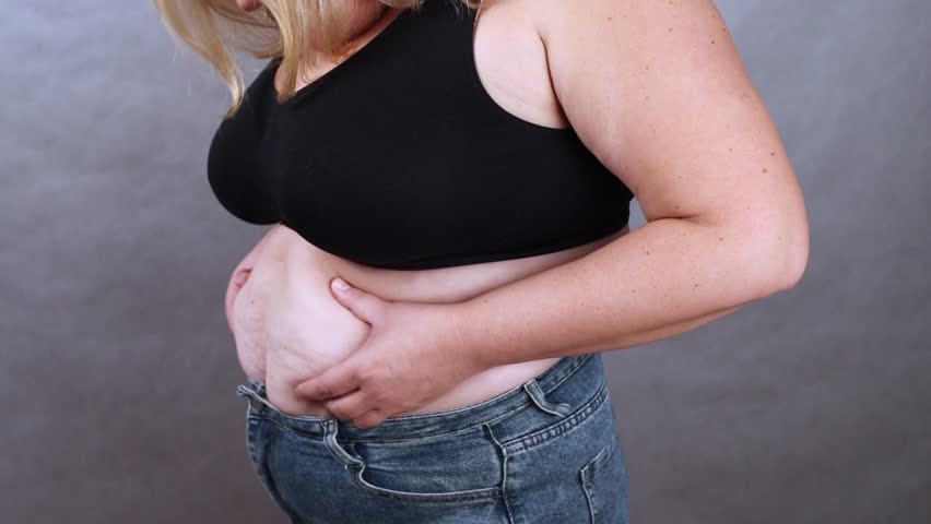 Woman in black top and jeans demonstrates body confidence by holding her midsection, showcasing a gradual progression of self-acceptance and body positivity in a studio setting