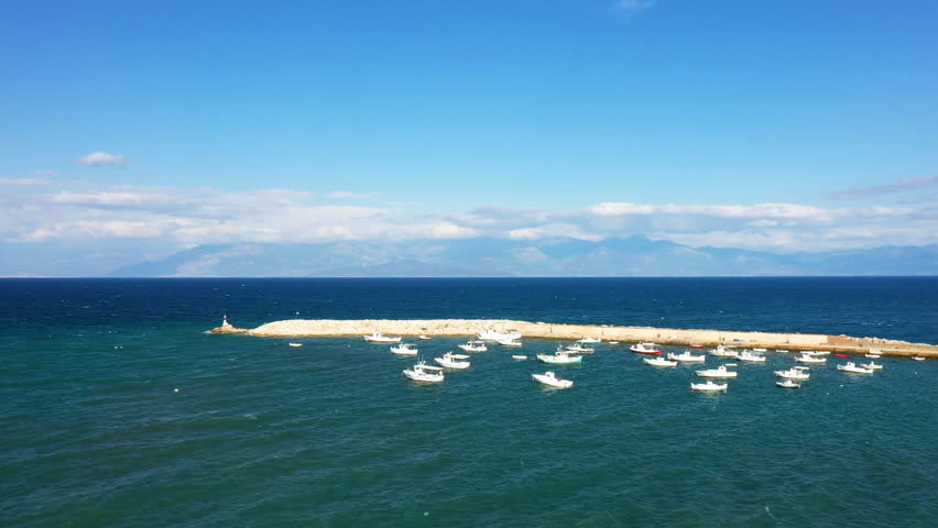 Aerial view of small boats anchored by a stone pier in the harbor of Koroni, Greece, with deep blue sea and mountains in the background under a clear sky.