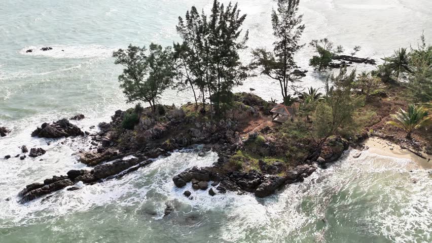 This aerial footage captures a stunning tropical beach paradise where green emerald ocean water surrounds a small rocky island and meets a quiet sandy coastline. Ocean waves move continuously across the shore, creating soft sea foam as they crash against natural rock formations and gently wash over the sand. The sunlight reflects beautifully on the surface of the sea, enhancing the fresh and calming atmosphere of this untouched tropical environment.

Tall coastal trees and tropical vegetation grow naturally along the island, adding depth and contrast to the rocky shoreline. From above, the composition reveals a perfect balance between water, land, and vegetation, presenting a peaceful yet dynamic coastal landscape. The movement of the waves brings life to the scene while maintaining a relaxing and cinematic mood.

This video is ideal for travel promotions, tourism marketing, luxury branding, environmental storytelling, cinematic backgrounds, wellness visuals, and nature documentaries. It represents freedom, serenity, and the timeless beauty of a tropical beach paradise, making it highly attractive for global stock footage buyers seeking authentic and naturally preserved coastal scenery.