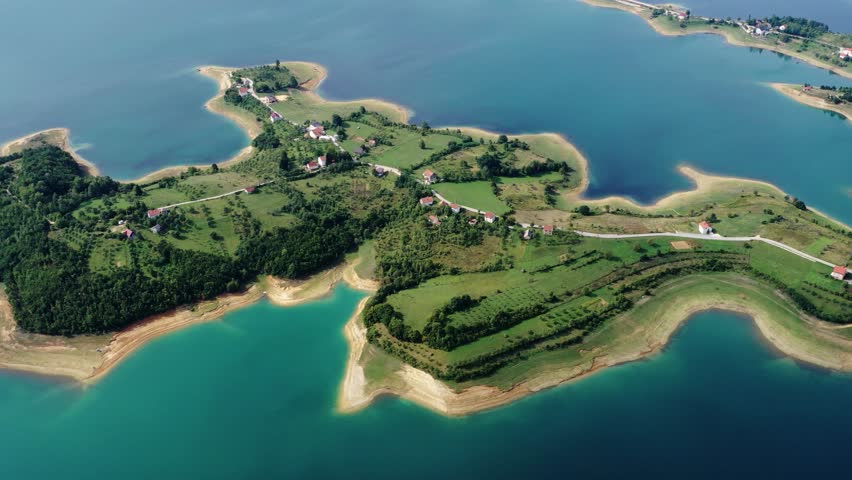 Aerial panoramic view of an island and curved lake shoreline in Bosnia and Herzegovina in 2019, showing green land, blue water and peaceful natural surroundings.