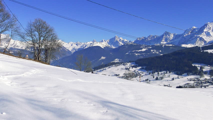 Scenic winter view of snowy fields, rustic barn, and the majestic Massif du Mont Blanc mountains under a clear blue sky. Idyllic alpine setting with traditional wooden chalet and distant village.