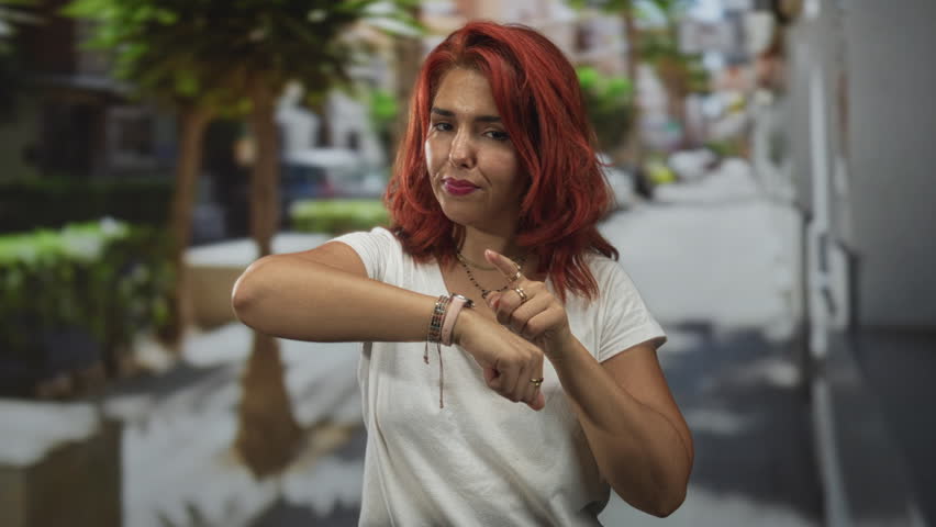 Woman with red hair checking watch on wrist and points finger to watch in city street lined with trees; running late impatience.