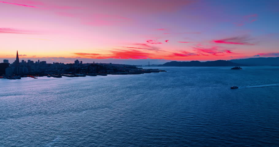 Boat moves by the blue waterscape of the bay. Dark skyline of San Francisco, CA, USA at sunset against pink sky at backdrop. Aerial view.