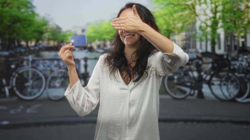 Woman holding creditcard while covering her eyes with one hand on a street lined with bicycles and a canal; privacy playful joyful.