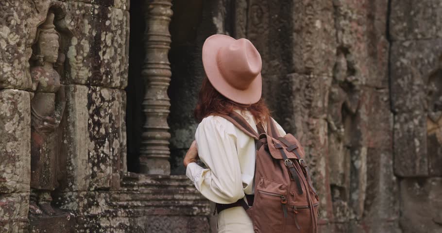 Stylish female traveler with hat and backpack explores ancient stone carvings at Ta Som temple, Angkor Wat, Cambodia. Conceptual lifestyle and tourism stock video for promo, ads and travel campaigns.