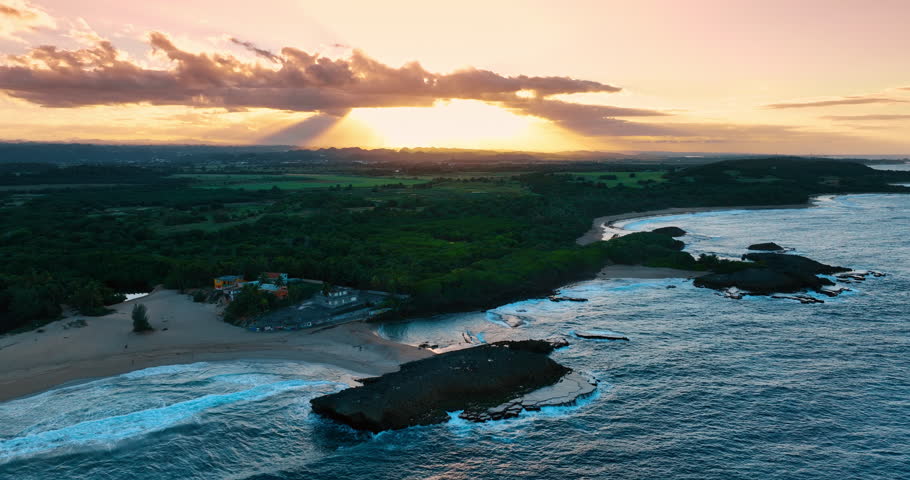 Footage over the coast with raging waves. Lush greenery covers the landscape. Clouds hide the setting sun in the sky. Puerto Rico.