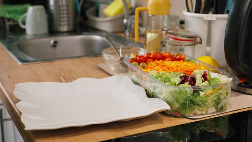 Woman hand use tongs to serve fresh, colorful vegetable salad from container onto white plate, showcasing healthy meal being prepared on kitchen countertop at home.