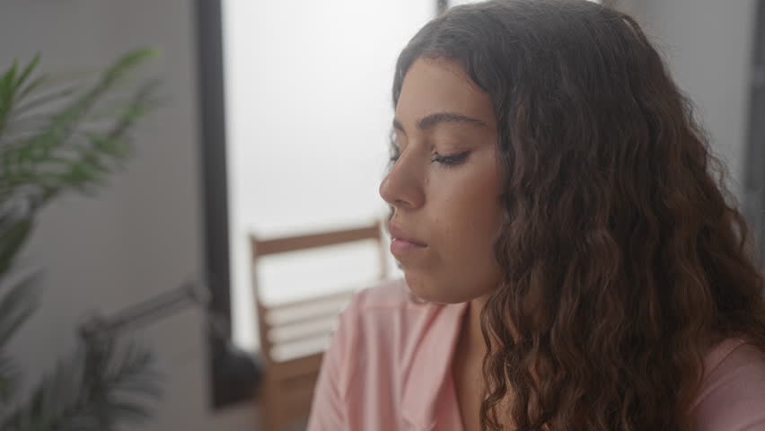 Hispanic woman with eyes closed in profile beside a houseplant and lamp in a studio, seated and softly backlit; reflection calm.