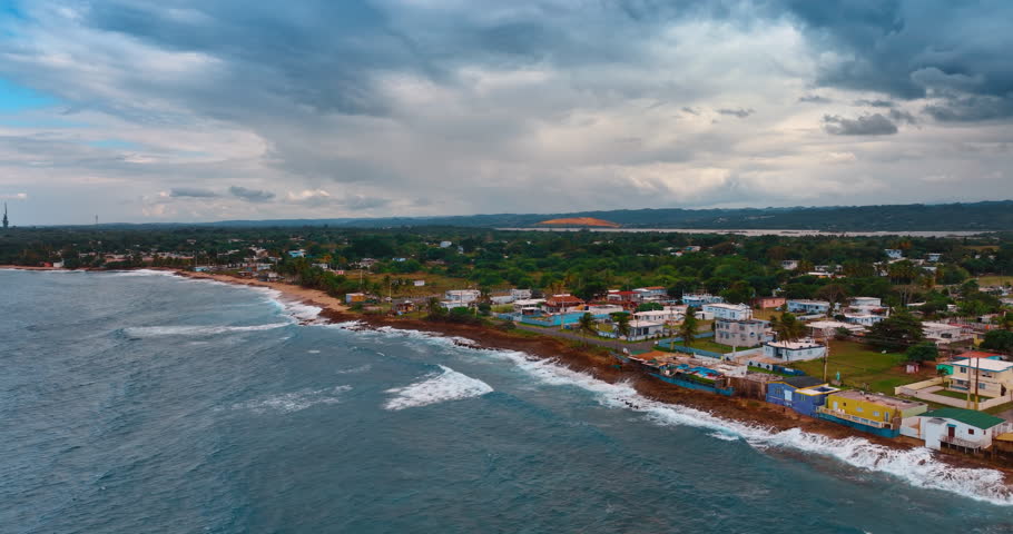 Raging waves hit the coast with row of buildings. Aerial perspective on the green cityscape in Puerto Rico under the grey cloudy sky.