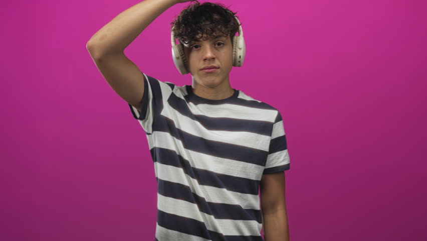 Teenager with headphones raises hand to head and gestures to chest against magenta studio wall; playfulness.