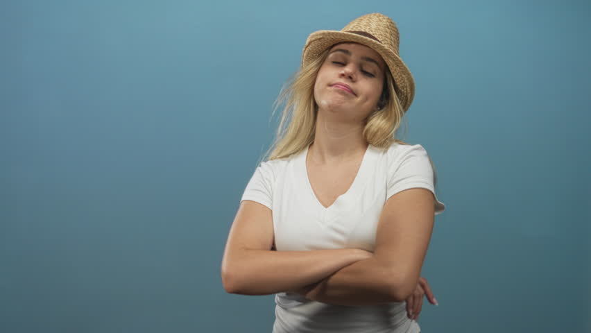 Young blonde woman wearing white vneck shirt and straw hat with crossed arms in blue studio; confidence.