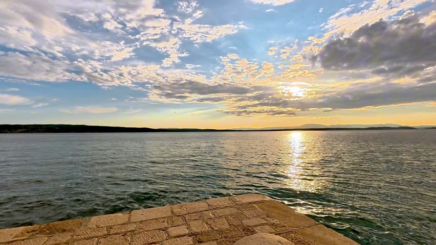 A sunset view over the Adriatic Sea from a stone pier in Crikvenica, Croatia. Golden hour light reflects on the water surface as the sun sets over the coastal horizon.