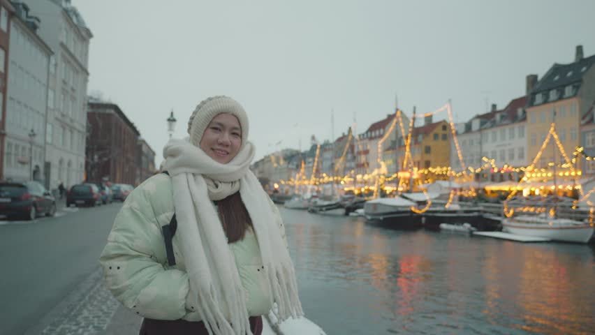 A serene winter view featuring a person wearing a warm coat and scarf near a festive waterfront, adorned with glowing lights and buildings reflecting in the water.