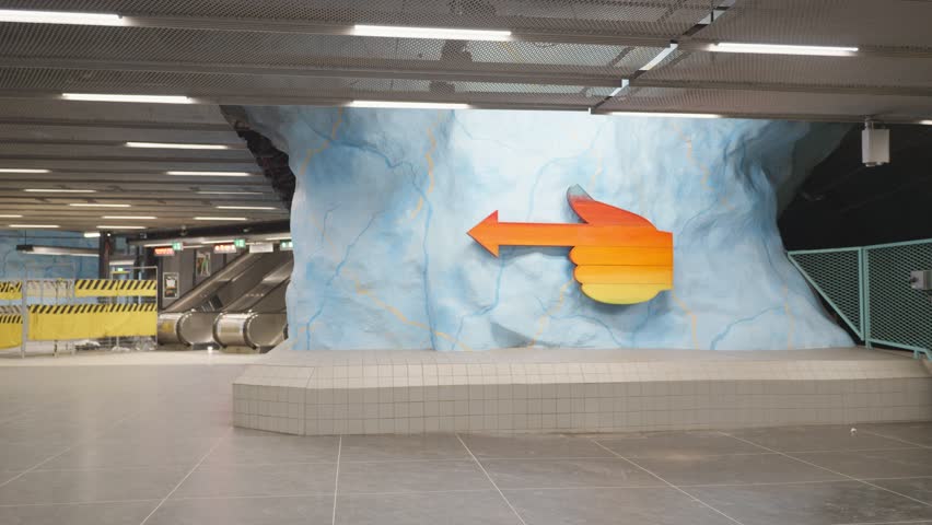 A colorful thumbs-up directional sign against a textured blue wall in a contemporary subway station. The artwork enhances the space, guiding passengers while adding a touch of creativity to public tra