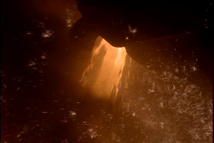 Steel works in Nizhny-Tagil, Russia. CU orange glowing molten iron pouring from ladle amid orange smoke, camera tilts down to rain of sparks hitting the floor.