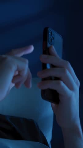 Exhausted young man alone in bed browses social media on his glowing phone at night, struggling with insomnia, stress and screen addiction until he becomes overwhelmed. Depression and doom scrolling 