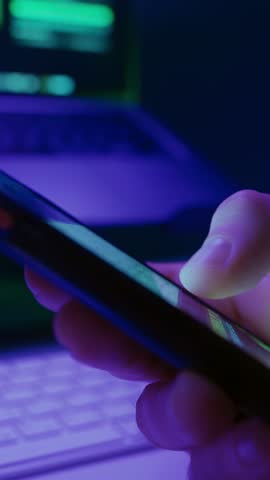 Close-up of persons hand scrolling on smartphone social media feed at night with laptop on background, illuminated by neon light. Digital addiction and modern online habits. Vertical video