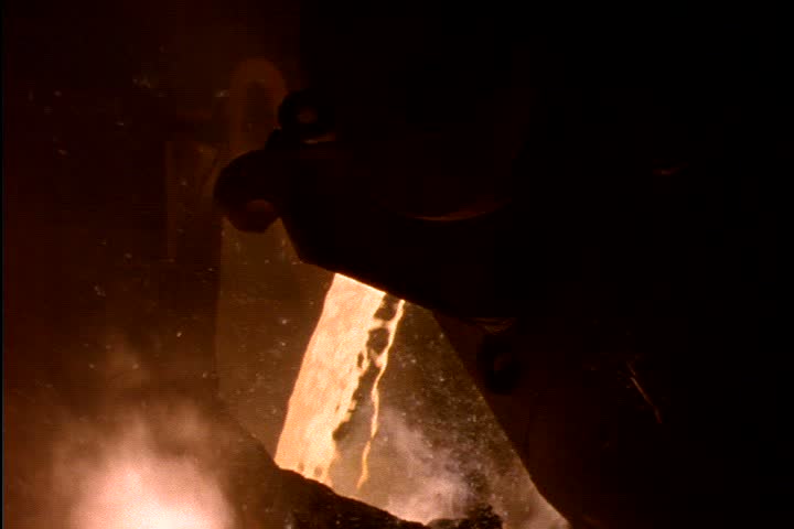 Steel works in Nizhny-Tagil, Russia. Dark silhouette of open hearth ladle in foreground, MS orange glowing molten iron pours from ladle in slow motion amid orange smoke and flames.