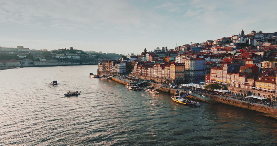Porto city historic Ribeira district vibrant riverside buildings and bustling promenade along sparkling Douro river with traditional boats floating under a clear sky. Aerial view drone flight footage
