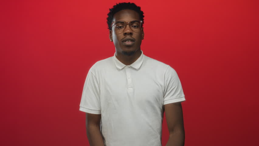 Man wearing glasses and white polo shirt making finger heart gesture in studio against red background; affection.