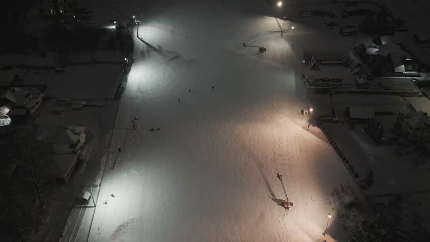 Aerial night approach toward snowy ski piste under artificial lights