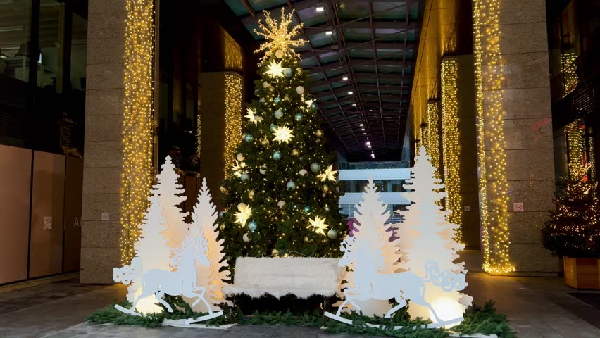 A festive interior view of a Christmas tree with gold and white decorations, surrounded by illuminated white decorative trees and a bench in a modern public space or lobby.