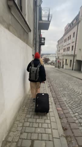 Traveler Pulling Suitcase Along Cobblestone Street, Red Beanie And Gray Backpack, Neutral Coat, Rolling Black Suitcase On Tiled Sidewalk, Narrow European Buildings With Satellite Dishes And Balconies