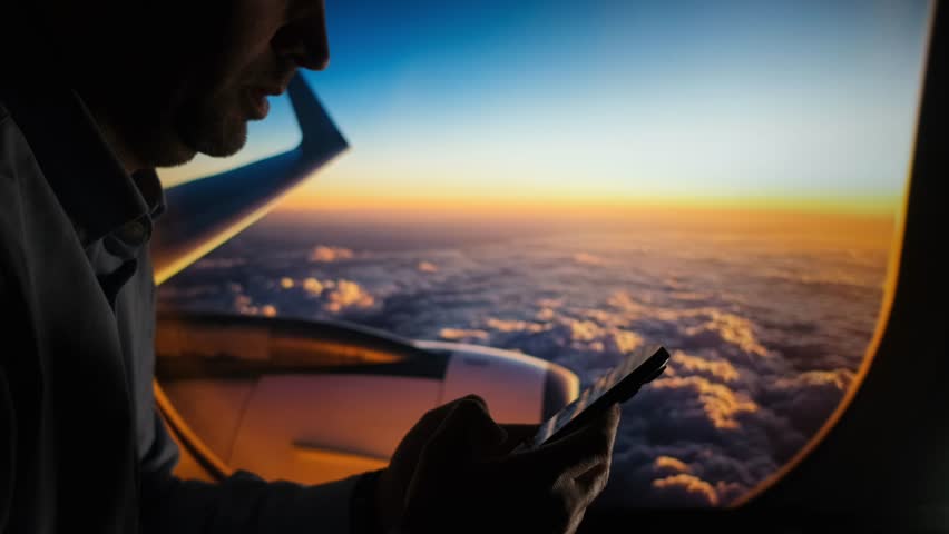 A man using a smartphone while flying in an airplane, silhouetted against a vibrant sunset sky above the clouds.