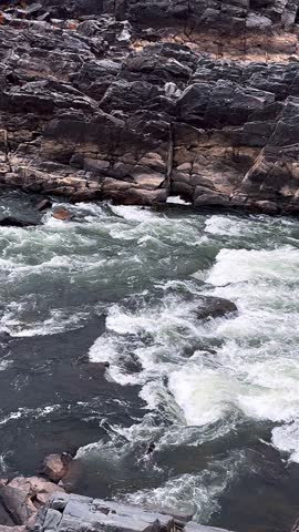 Fast moving white water surges through a narrow river channel, crashing over dark stone formations and creating strong currents and splashing foam.
