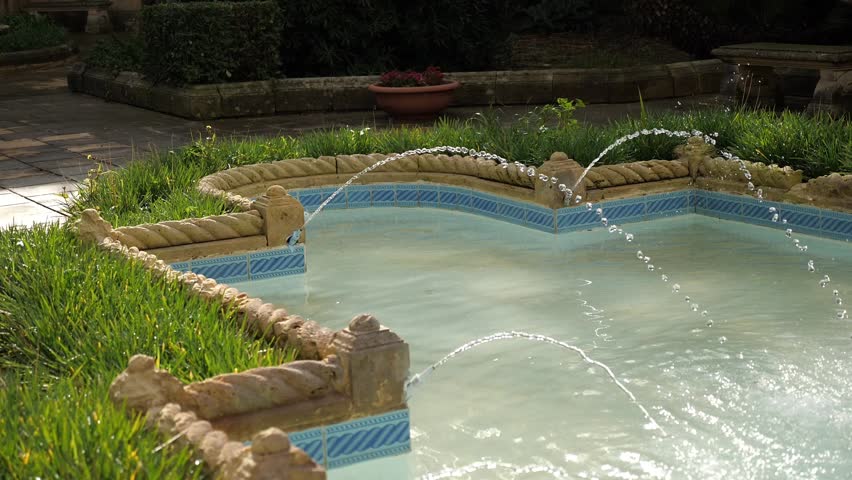 Close up tiled garden fountain with water jets and green plants under bright sun. 
