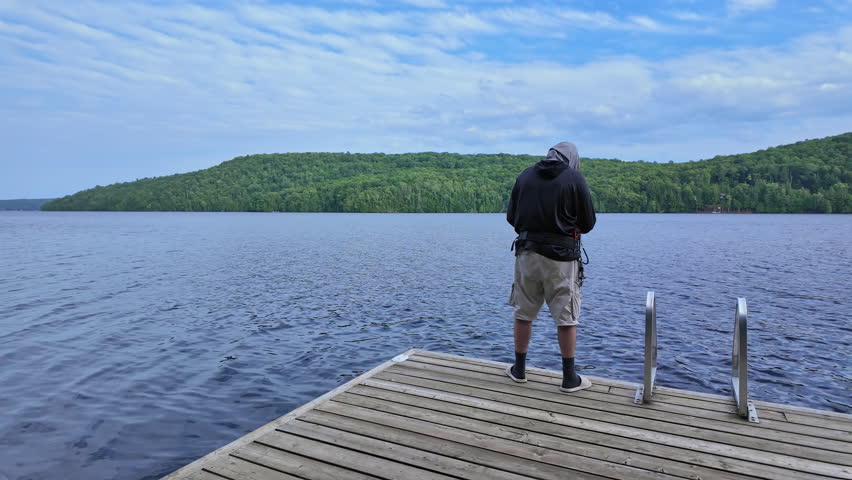 Man fishing at serene lake from the boat dock, casting fishing spinning rod and using reel. Sport angler using UV protective head hood or balaclava neck gaiter as sun mask. Fishing summer vacation