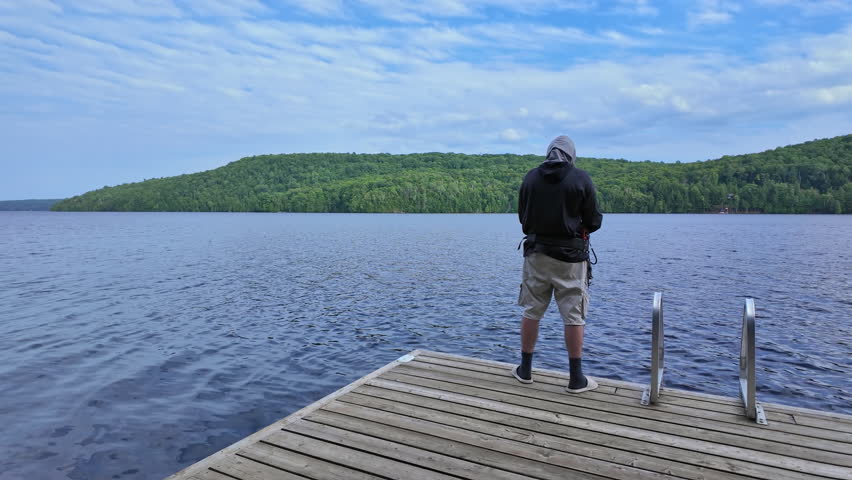 Man fishing at serene lake from the boat dock, casting fishing spinning rod and using reel. Sport angler using UV protective head hood or balaclava neck gaiter as sun mask. Fishing summer vacation