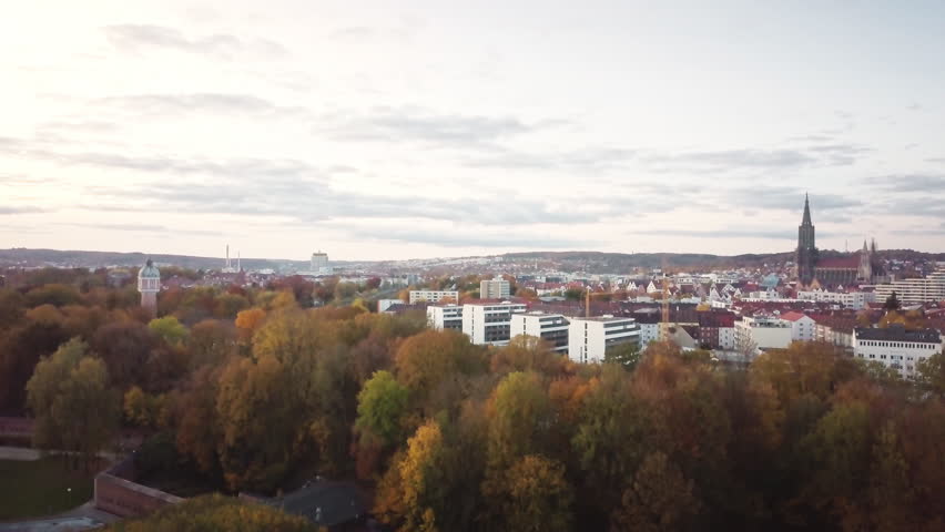 4K aerial drone rising over Ulm cityscape with autumn forest. Premium footage revealing city skyline, cathedral spire, and vibrant fall colors. Perfect for travel, documentaries, and corporate content