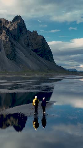 Two adventurers explore the breathtaking landscape of Stokksnes, with Vestrahorns jagged peaks reflected in calm waters. A picturesque moment in Icelands natural beauty unfolds under a vibrant sky.
