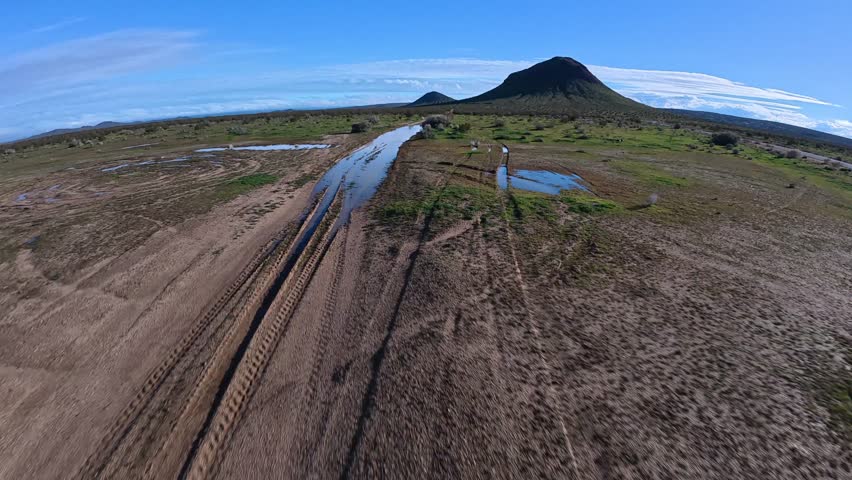 A drone follows a sandy desert road with rain puddles after rainfall in California. As the flight rises, a large butte appears, revealing the vast, green Mojave landscape under blue skies.