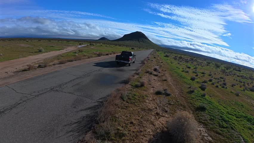 A drone tracks a pickup truck moving through the Mojave Desert in California. The flat landscape is mostly green with wet patches, and a large butte dominates the horizon beneath clear skies.