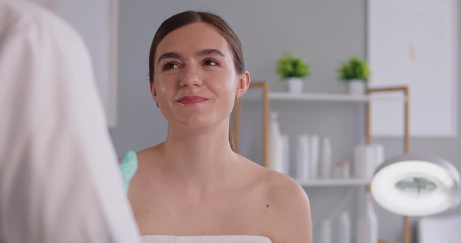 Esthetician wearing green gloves prepares for facial treatment in a bright clinic, assessing skin and client comfort. Cosmetologist gently examines the woman's face. Calm skincare scene.