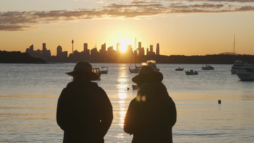 Silhouette of two people wearing hats standing by the water in Watsons Bay, watching the sun set directly behind the Sydney city skyline.