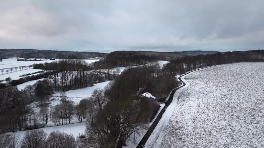 Peaceful snowy forest landscape with street and leafless trees on cloudy winter day. Grey sky and snow-covered farm fields in America. Aerial wide shot. No people on path.