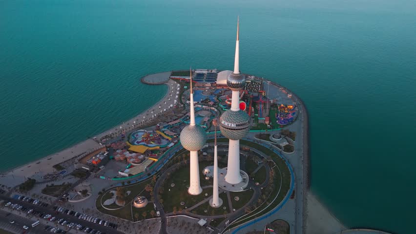 Wide drone aerial of Kuwait Towers at blue hour, surrounded by waterfront park, promenade and seaside amusement park on the Arabian Gulf—clean ocean backdrop, iconic Kuwait City landmark.
