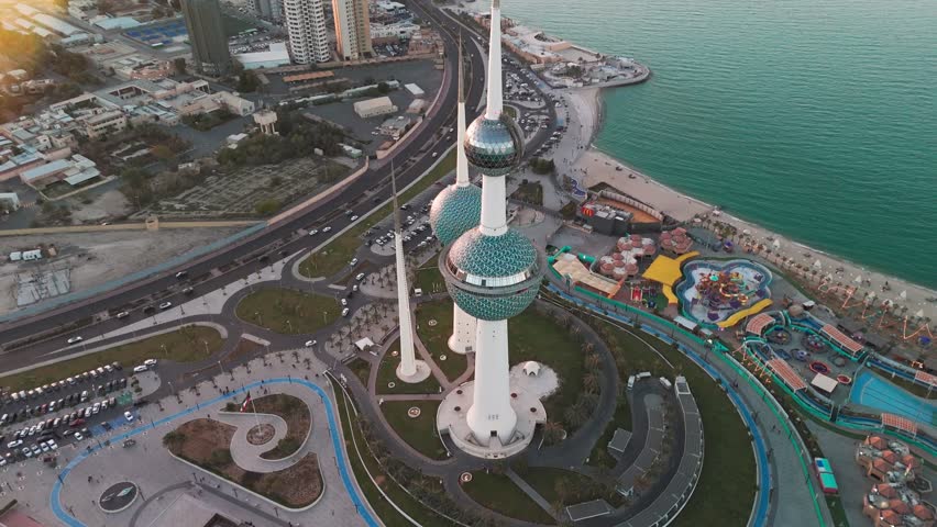 Top-down drone view of Kuwait Towers with turquoise mosaic spheres, coastal roads, beachfront and waterfront park beside the Arabian Gulf at golden hour—ideal tourism, landmark, city travel footage.
