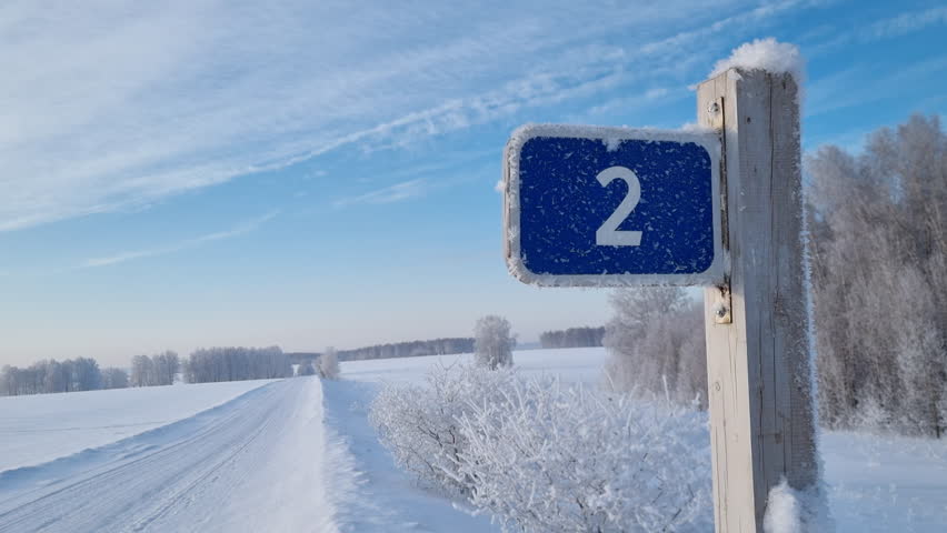 Blue number two sign covered with hoarfrost on a wooden post. A beautiful, remote country road is blanketed in fresh, deep snow